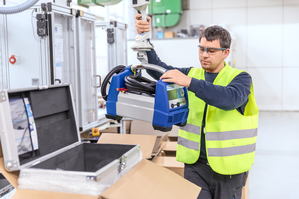 Het handvat van het FRIAMAT lasapparaat past perfect op de haak van de JumboFlex slangheffer. Zo tilt de werknemer het apparaat in het teststation en vervolgens in de koffer.